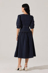 A woman is wearing a navy blue dress with puffed three-quarter-length sleeves, a fitted waist with white topstitching details, and a gathered, full-length skirt. She pairs the dress with light-colored, high-heeled sandals that have ankle straps.